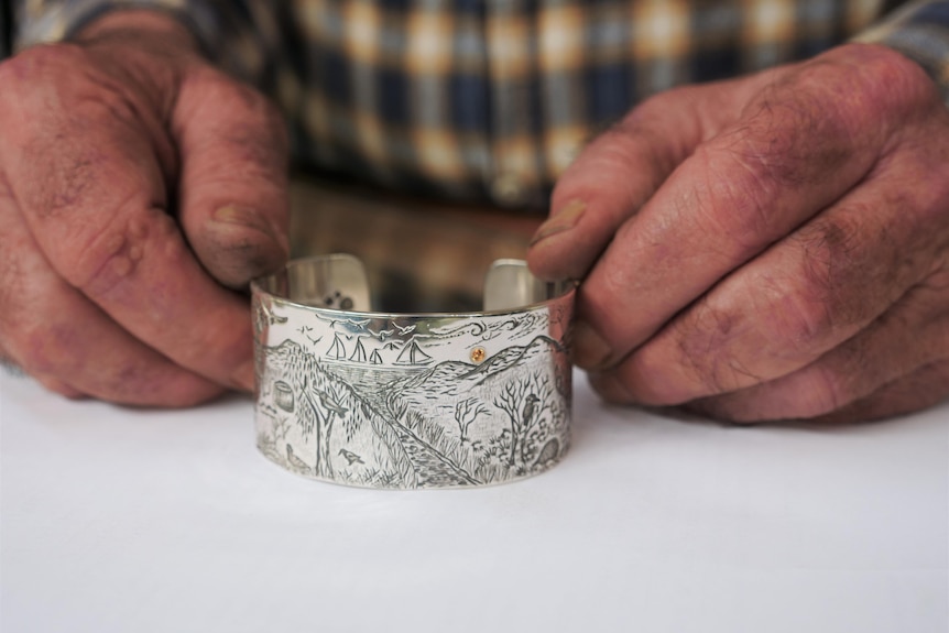 A man's hands holding a silver cuff with a landscape stamped into the metal. 