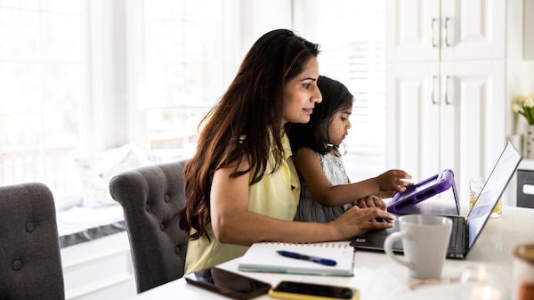 A woman and daughter sit side by side at a dining table, working on a laptop and tablet.