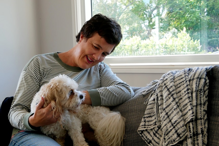 A woman with short brown hair pats a small white dog on her lap