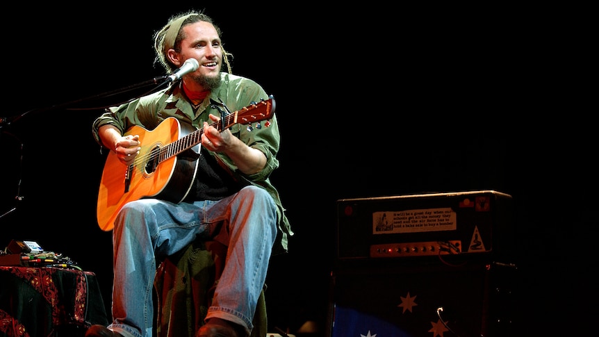 John Butler on stage playing guitar and smiling. He wears a green headband and green shirt.