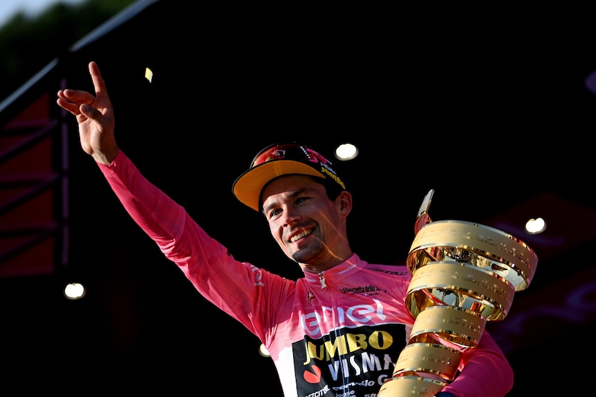 A cyclist in a pink jersey points in celebration while he holds the trophy after winning the Giro d'Italia.