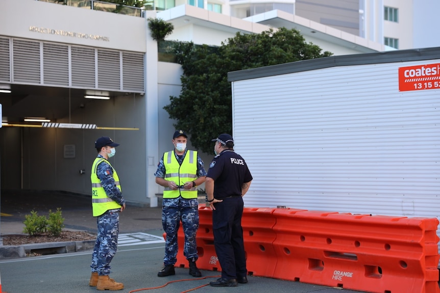 Two in military outfits and yellow high-vis vests talking to a police near an orange barricade