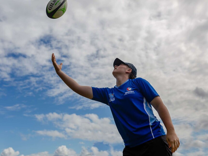 A girl throws a rugby ball in the air. Only blue sky and clouds visible in background.