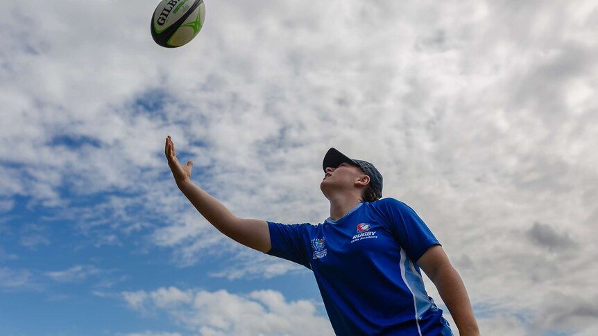 A girl throws a rugby ball in the air. Only blue sky and clouds visible in background.