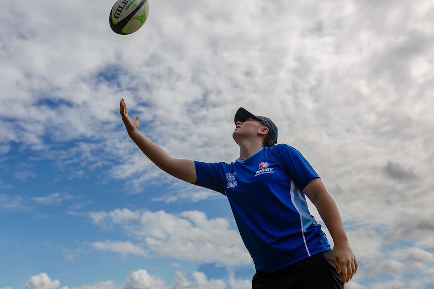 A girl throws a rugby ball in the air. Only blue sky and clouds visible in background.