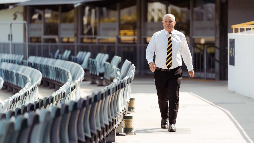 A wide shot showing Terry Waldron walking around a terrace of the WACA Ground in a white shirt, black pants and tie.