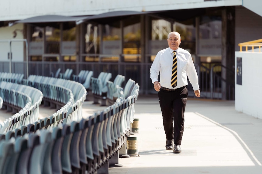 A wide shot showing Terry Waldron walking around a terrace of the WACA Ground in a white shirt, black pants and tie.