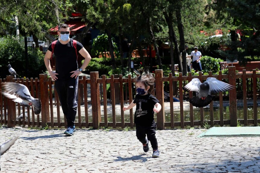 a child wearing a face mask chases after pidgeons as a man with a face mask and sunglasses watches on