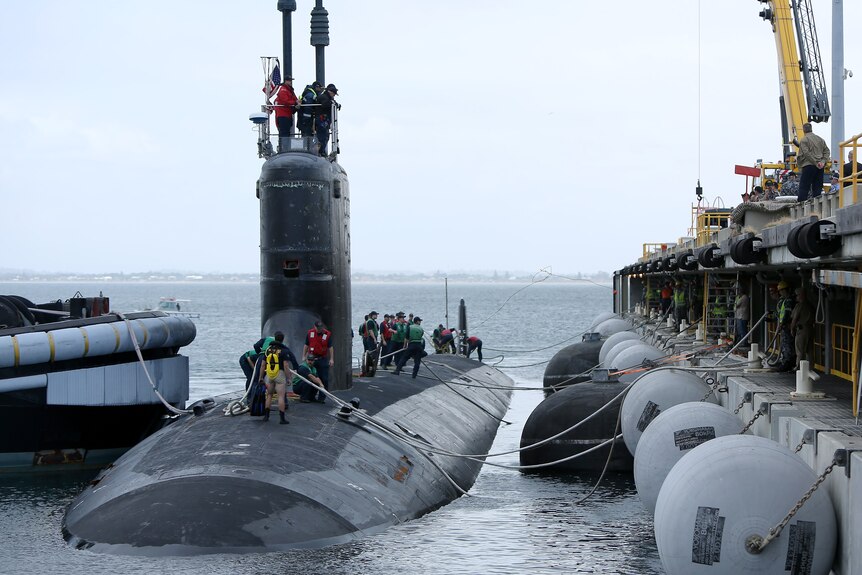 A dark grey submarine floats on the ocean surface beside a dock as people stand on top of it.