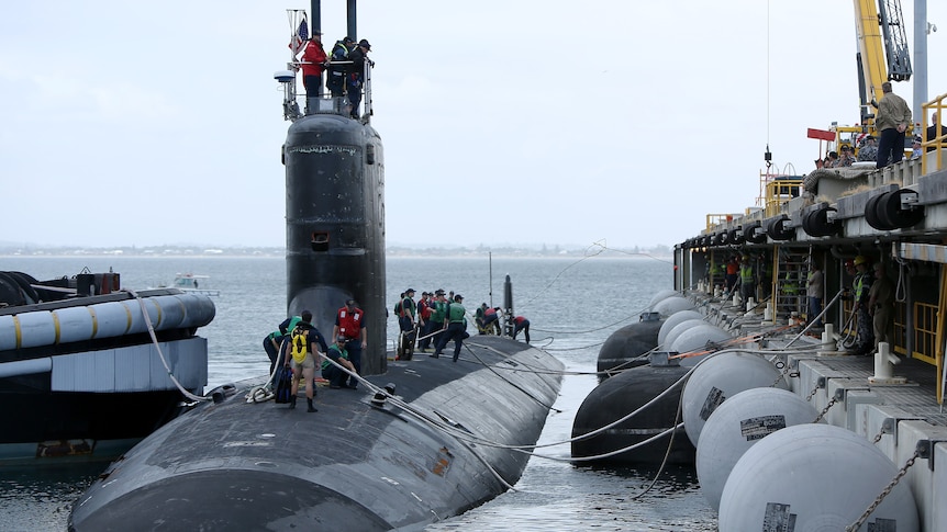 A dark grey submarine floats on the ocean surface beside a dock as people stand on top of it.