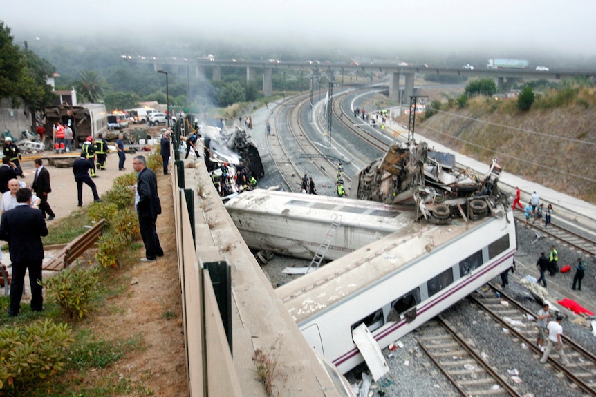 A train below crashed into a cement wall, derailed and on its side. People above are watching as rescue workers recover victims.