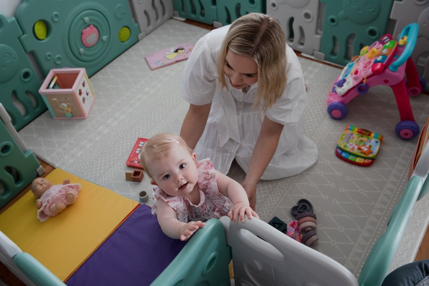 A woman playing with her bub, who's standing up and holding onto a cot.
