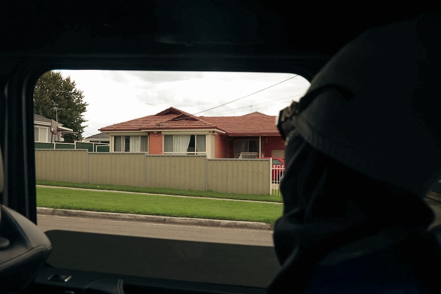 A person wearing a beanie, mask and sunglasses looks out the window of a car at a suburban house.