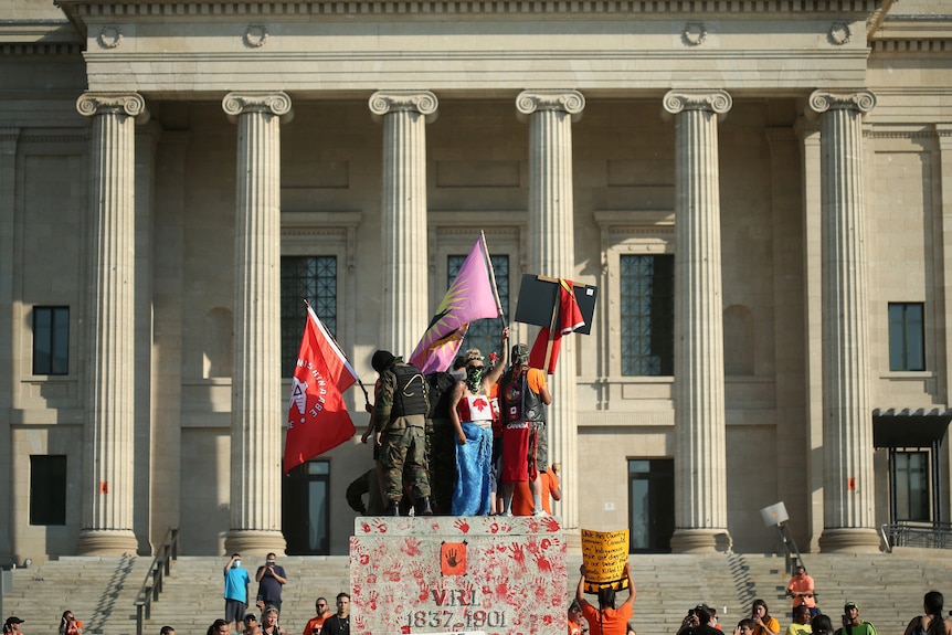 A group of protestors with First Nation flags and raised fists gather before a government building 