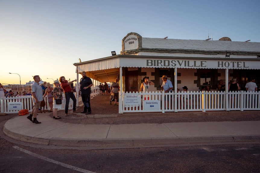 Pub with white picket fence, people standing out in front, sunset in the background.