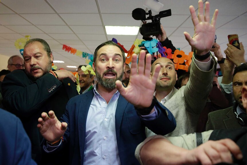 Santiago Abascal with his mouth open and his hand up surrounded by people and cameras after casting his ballot.