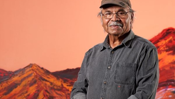 aboriginal man in baseball cap and glasses stands with hands clasped in front of large painting featuring orange sky & red hills