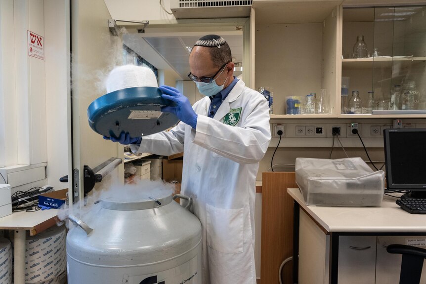 A man wearing a white lab coat, gloves, mask and yarmulke lifts the lid off a vat in a science lab