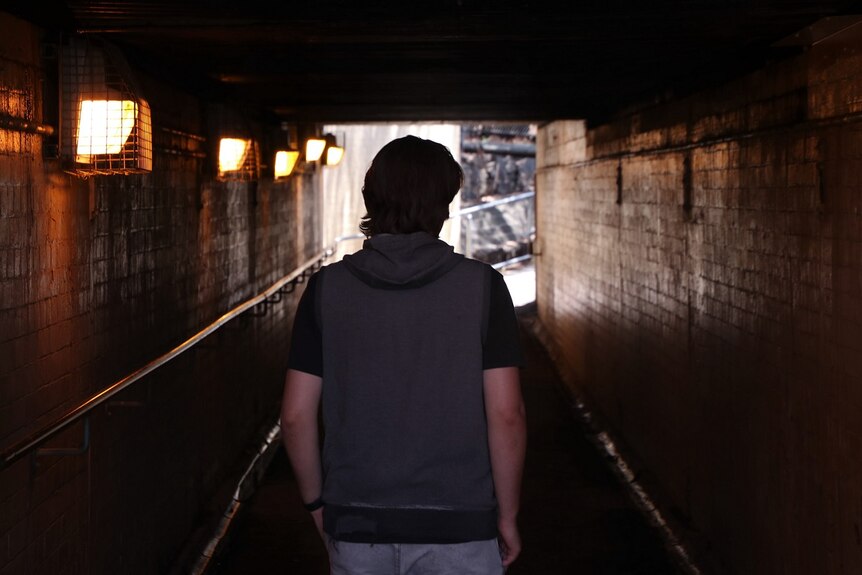 Allan walks away from the camera through a tunnel, with lights down one wall.