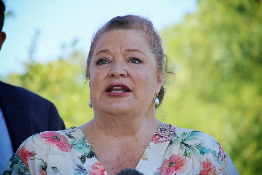 A close up of Sue Ellery standing outside with green trees and blue sky in the background, wearing a floral shirt.