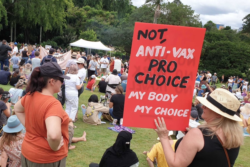 Protesters gather in a park where a woman is holding a red sign reading "Not anti-wax, pro choice, my body my choice"