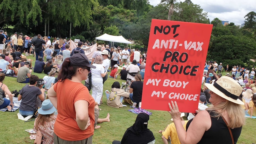 Protesters gather in a park, with one woman holding a red sign reading "Not anti-vax, pro choice, my body my choice"