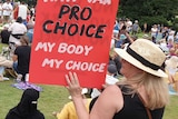 Protesters gather in a park, with one woman holding a red sign reading "Not anti-vax, pro choice, my body my choice"