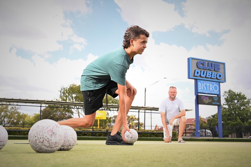 A teenage boy rolling a ball on a bowling green while a man watches on.