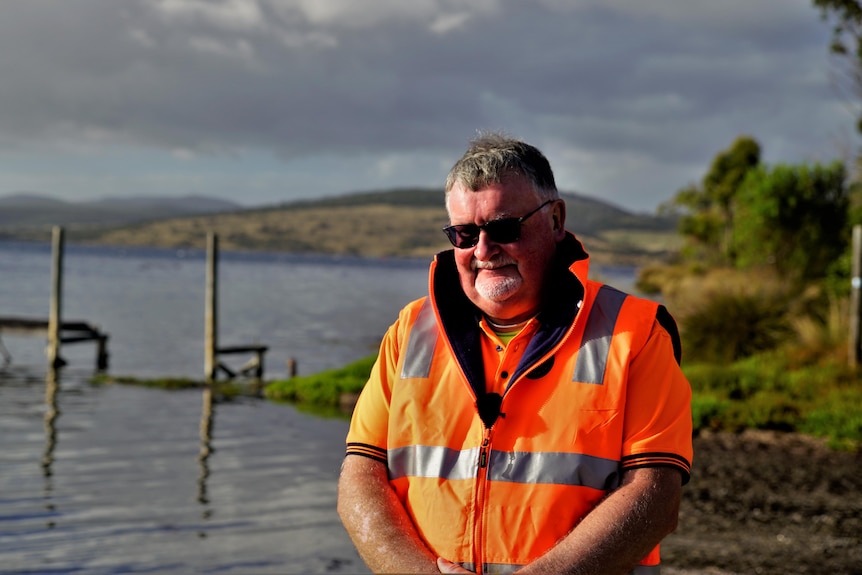 Man wearing sunglasses and hi vis orange vest stands in front of oyster growing area.