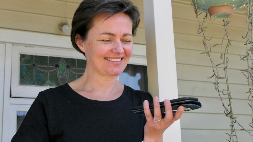 A woman stands out the front of a house, smiling and holding a phone.