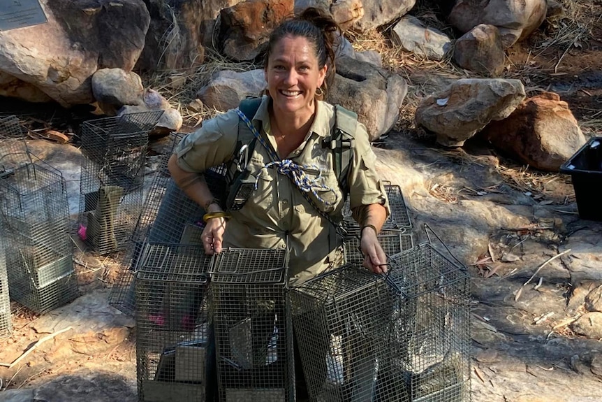 A smiling woman in khaki holds several metal traps in front of a rocky background, dark hair tied up.