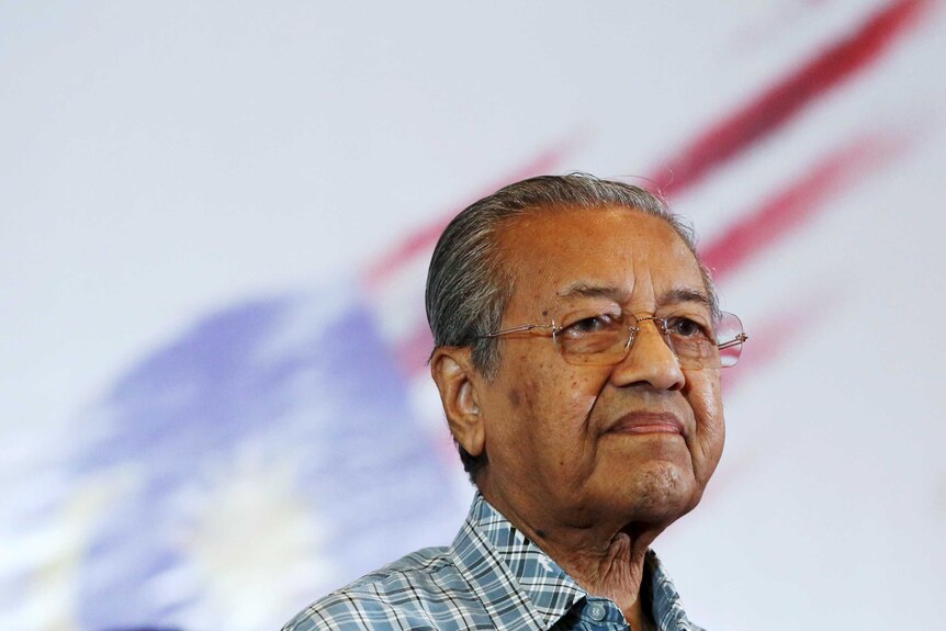 Mahathir Mohamad stands in front of flag looking into distance