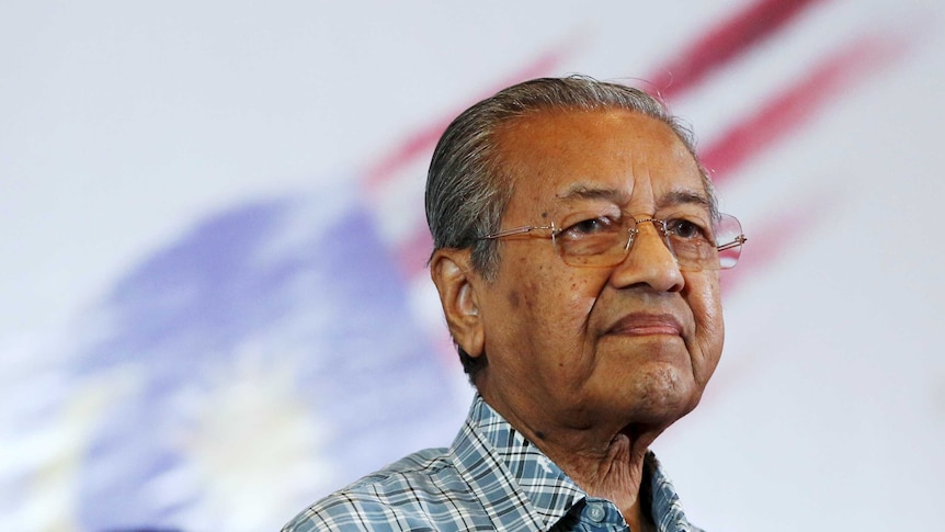 Mahathir Mohamad stands in front of flag looking into distance