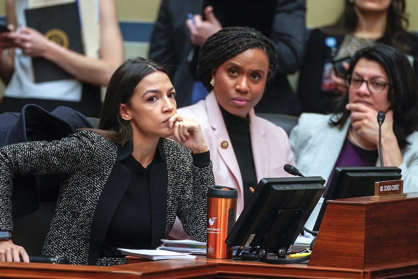 Behind redwood desks, Alexandria Ocasio-Cortez, Ayanna Pressley, and Rashida Tlaib listen behind small monitors and mics.