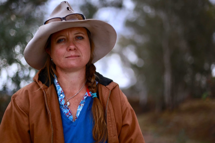 A woman wearing a brown jacket and Akubra hat, standing in bushland looking unimpressed.