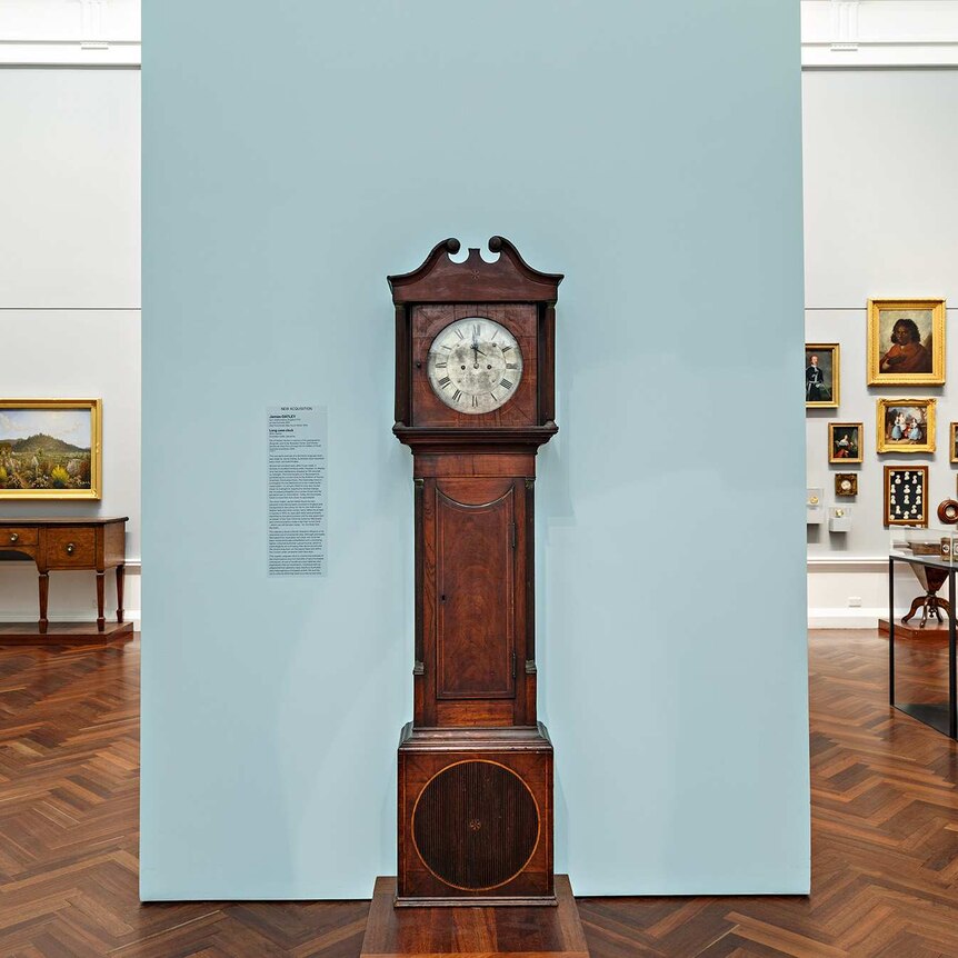 A large standing clock made of dark brown wood with a white face, in front of a light grey wall