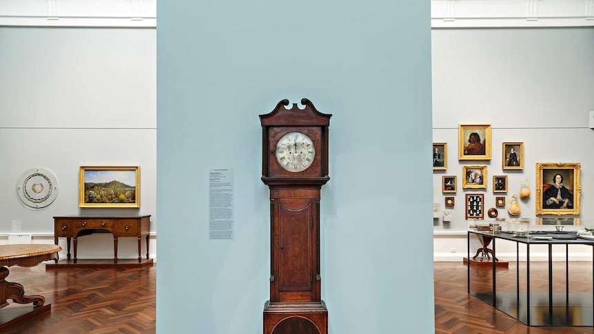 A large standing clock made of dark brown wood with a white face, in front of a light grey wall