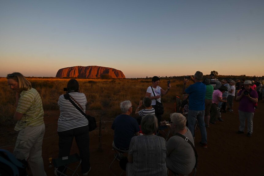 Crowds of tourists take photos of Uluru from a distance behind a fence.