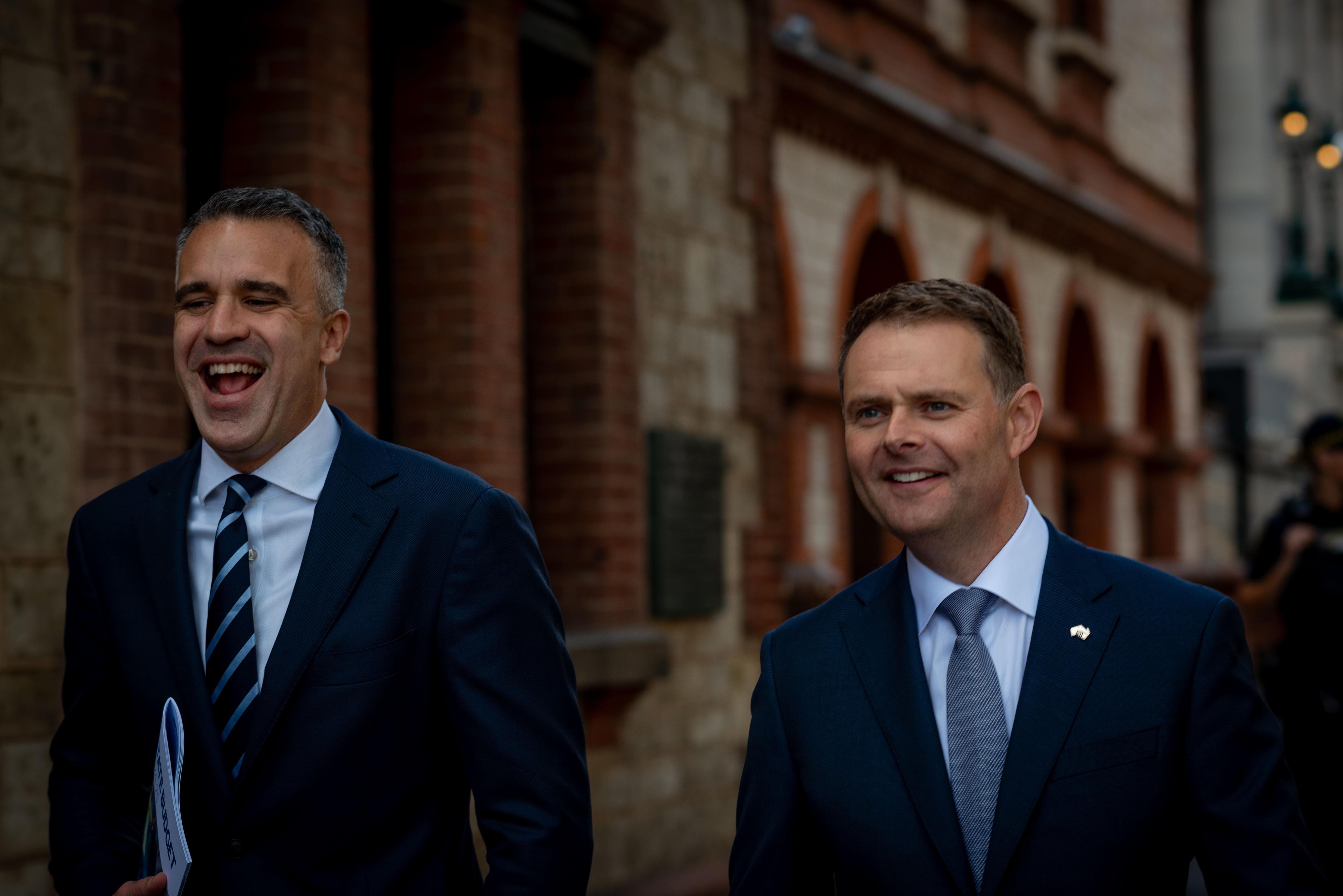 Dois homens de terno escuro e gravata azul caminham por North Terrace.