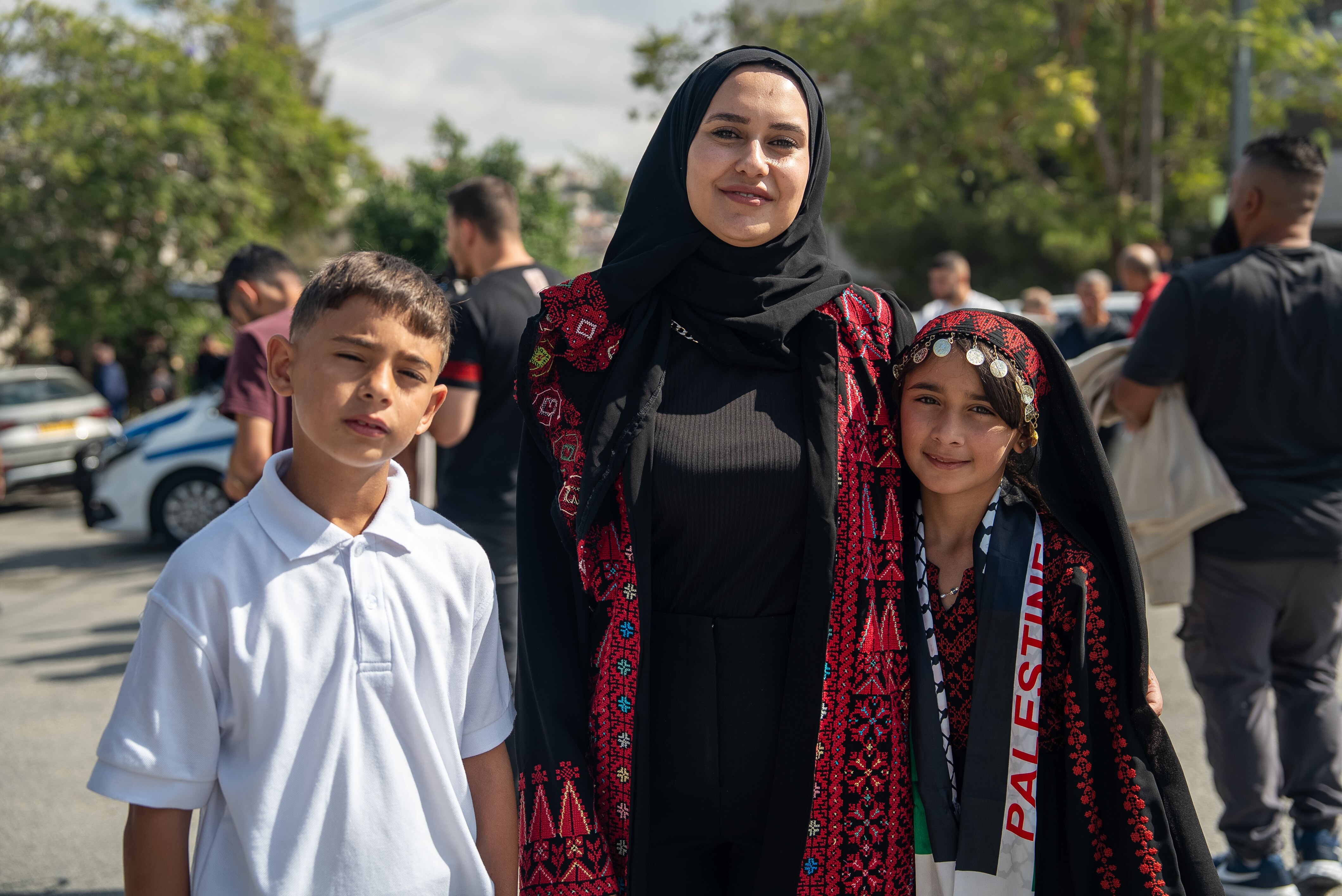 A woman wearing traditional Palestinian dress smiles with her son and daughter on a street.