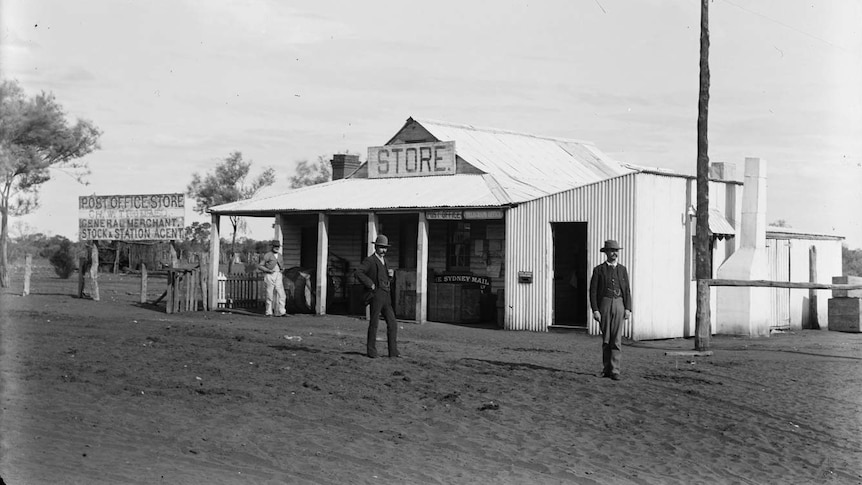 A black and white photograph of a corrugated iron building with a sign reading 'store'. Three men with suits and hats nearby