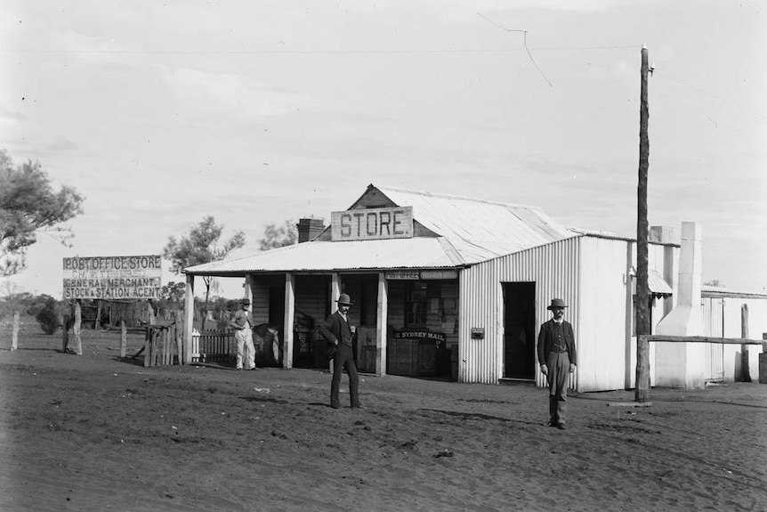 A black and white photograph of a corrugated iron building with a sign reading 'store'. Three men with suits and hats nearby