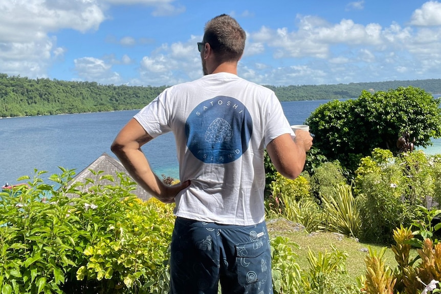 A man wearing a white tee standing looking out over lush greenery and blue water