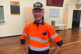 A man standing in a town hall, wearing an orange high-vis shirt with a black cap.