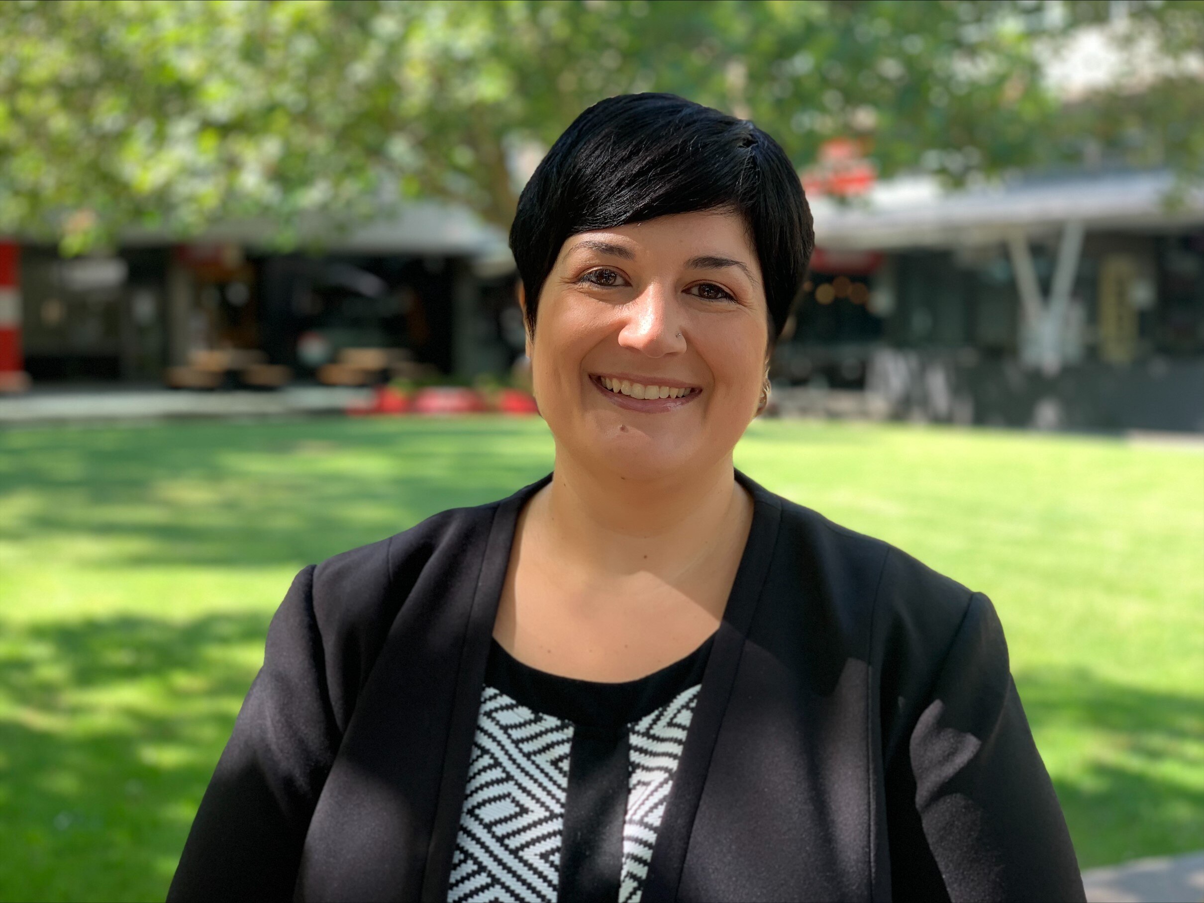 A woman with short black hair, and black and white striped shirt and black blazer stands in a grassy courtyard and smiles.