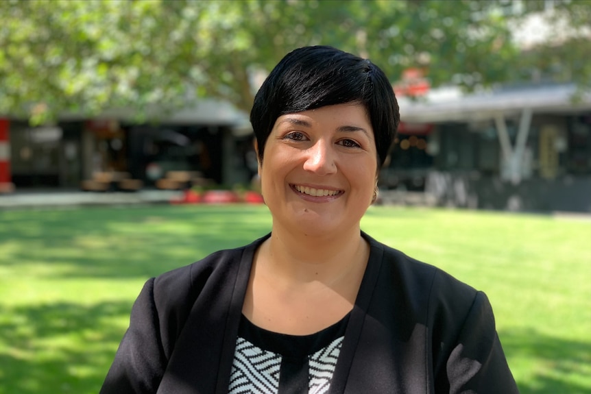 A short black-haired woman, a black and white striped shirt and a black blazer standing in a lawn courtyard and smiling.