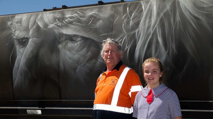 An older man and school girl standing in front of a portrait of a face painted on a train carriage