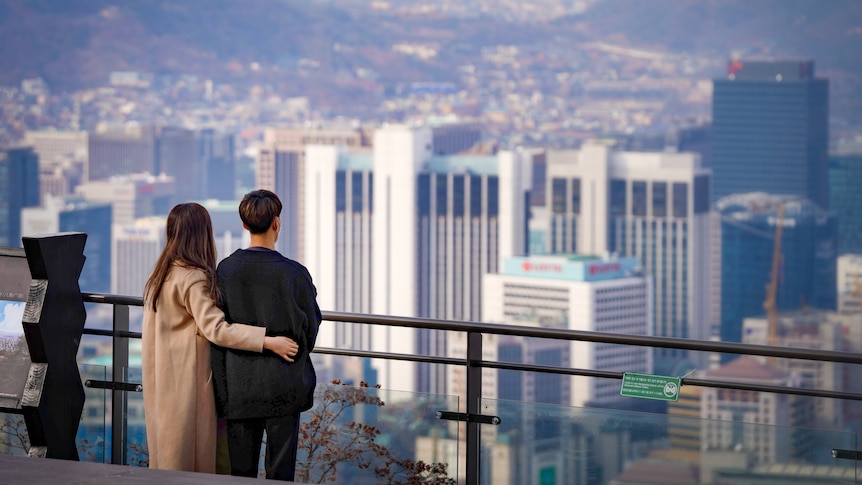 A Korean woman puts her arm around a man as they look across a railing at the Seoul skyline