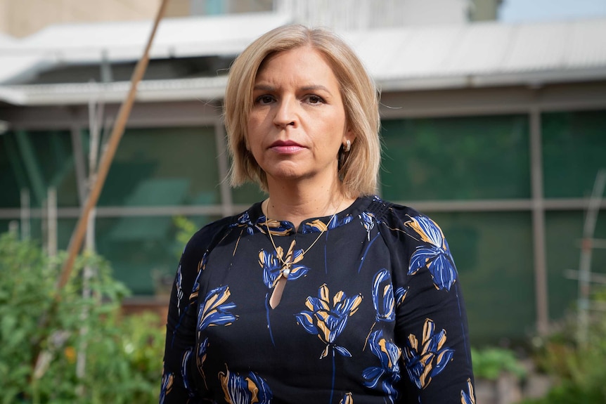 A woman with shoulder-length blonde hair, wearing a dark blouse with a floral design, stands outside looking sombre.