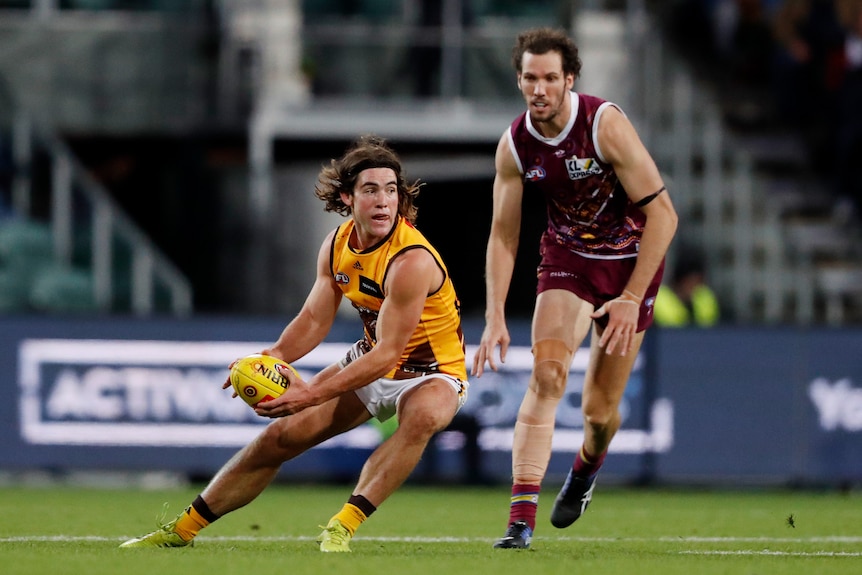 A Hawthorn AFL player holds the ball as he props to change direction with a Brisbane defender watching behind him.
